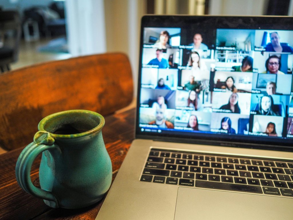 Una computadora portátil que muestra una videoconferencia con los rostros de varios participantes está sobre una mesa de madera junto a una taza de cerámica verde, sugiriendo una reunión virtual.