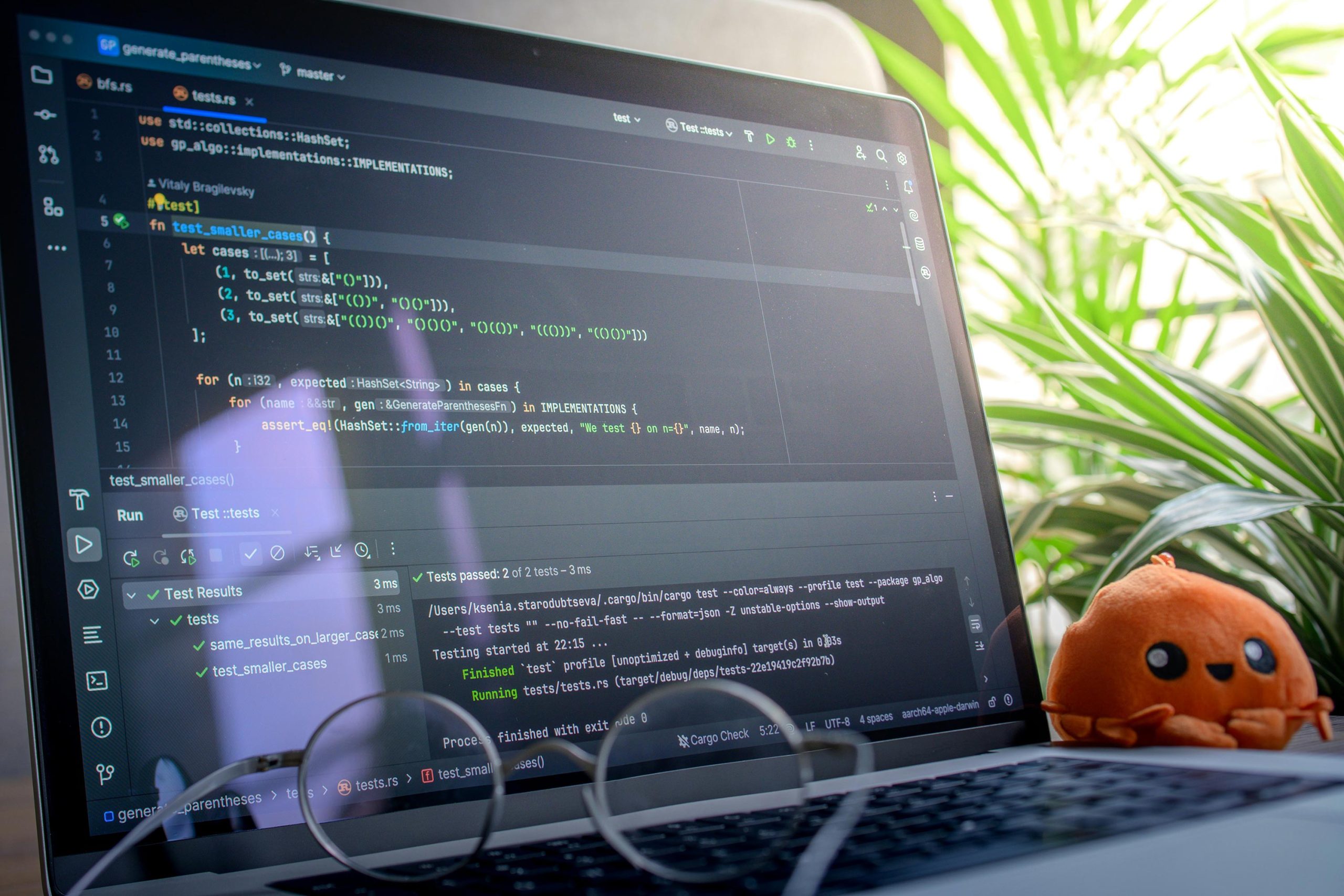 Una computadora portátil que muestra código en un entorno de desarrollo con una planta frondosa y un peluche naranja al fondo. La escena es tranquila y enfocada.