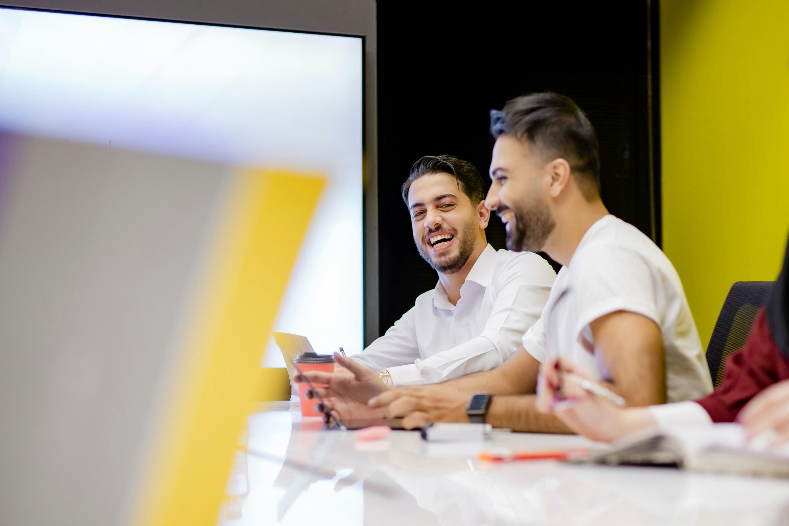 Dos hombres están sentados en una mesa de conferencias, sonriendo y conversando. El fondo presenta una pared de color amarillo brillante, creando una atmósfera alegre y animada.